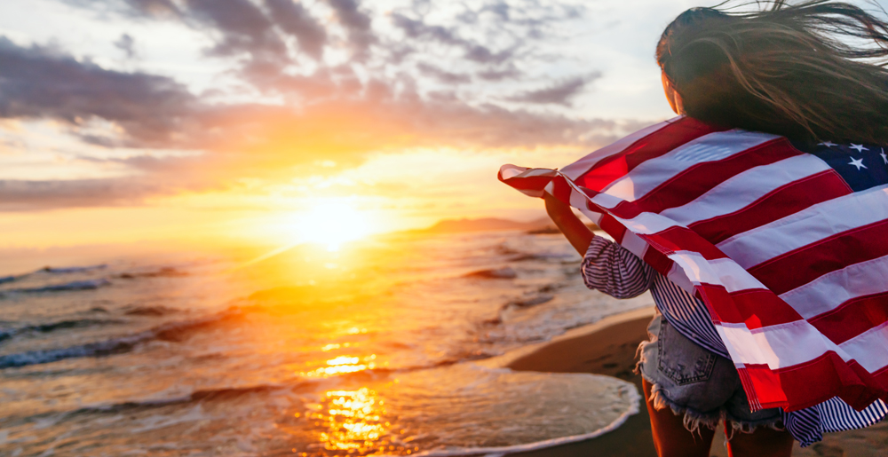 Person on a beach wearing an American flag as a cape at sunset, waves lapping the shore behind them.