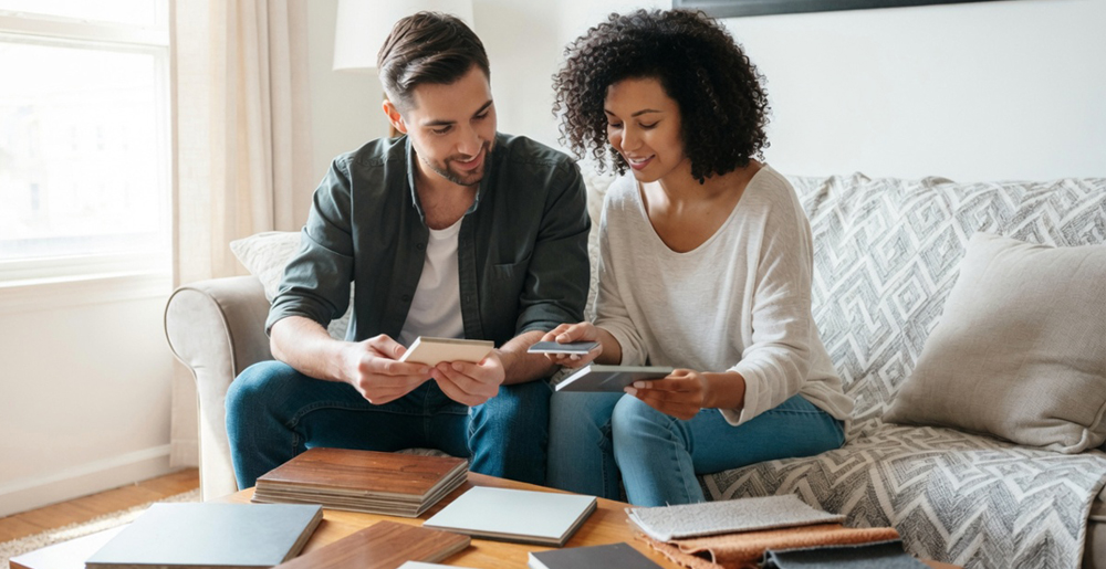 Couple sitting on a sofa, comparing tablets and color swatches for home decor.